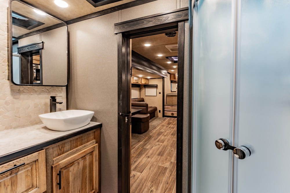 RV bathroom vanity with a white vessel sink and tiled backsplash, open doorway to living area with brown sofa and wood cabinets in background