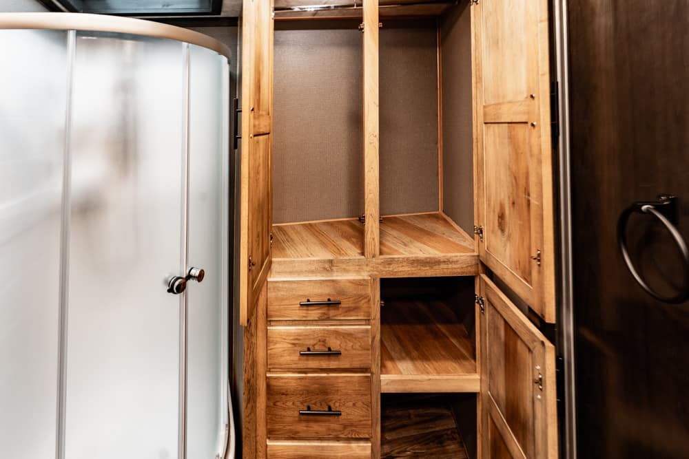 Wooden bathroom cabinet with open shelving and four drawers, doors open showing storage space between a curved shower and dark wall paneling.