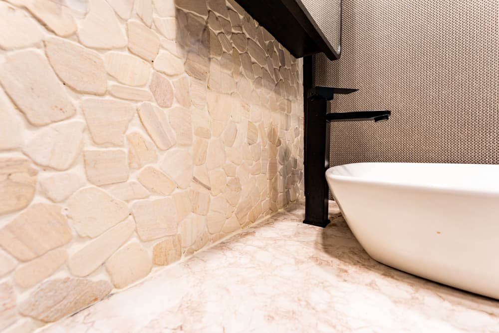 Modern bathroom sink with a tall black faucet, white vessel basin, beige stone-tile wall, and marble countertop.