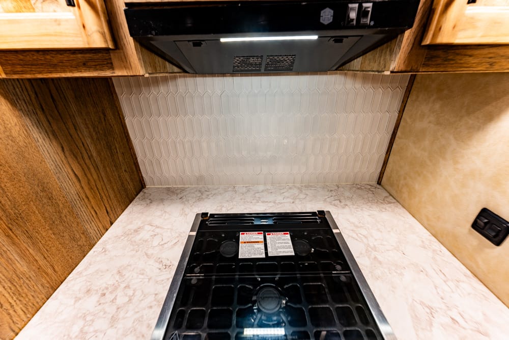 Compact kitchen stove with a black glass cooktop set into a marble-looking counter, range hood above, wood cabinets, and a light tile backsplash.