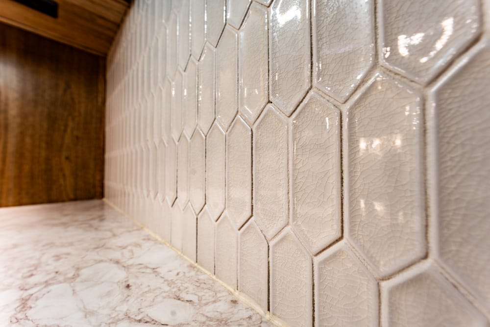 Interior view of a wall covered in glossy beige hexagonal tiles with visible crackle glaze, meeting a white marble floor; a wooden panel is visible at the far end.