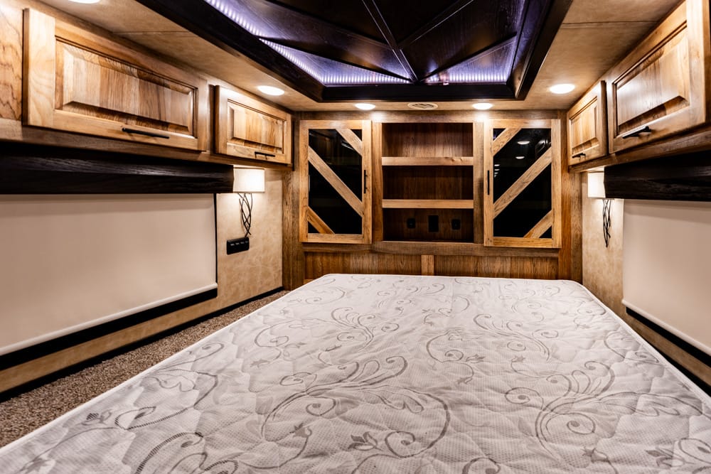 Wood-panelled RV bedroom with a large bed, patterned quilt, and built-in bedside cabinets and lighting above.