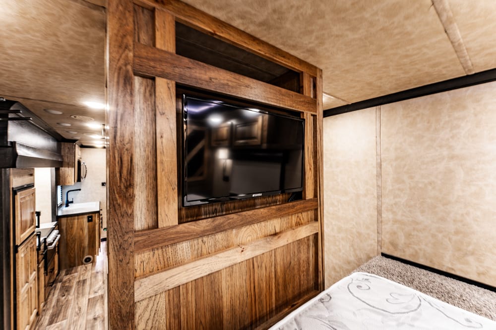 Rustic wood-paneled interior with a mounted flat-screen TV, kitchen area to the left and a bed at right in a camper/RV-like space.