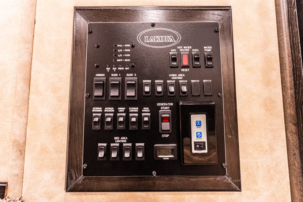 Close-up of a Dakota-brand RV electrical control panel with many labeled switches for awning, lighting, slides, and generator start/stop, mounted in a beige wall frame.