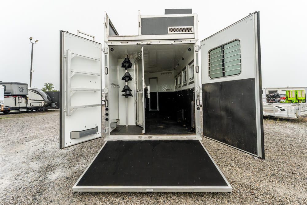 Open horse trailer with rear ramp down, showing an empty interior with stalls and storage on the left, parked on a gravel lot.