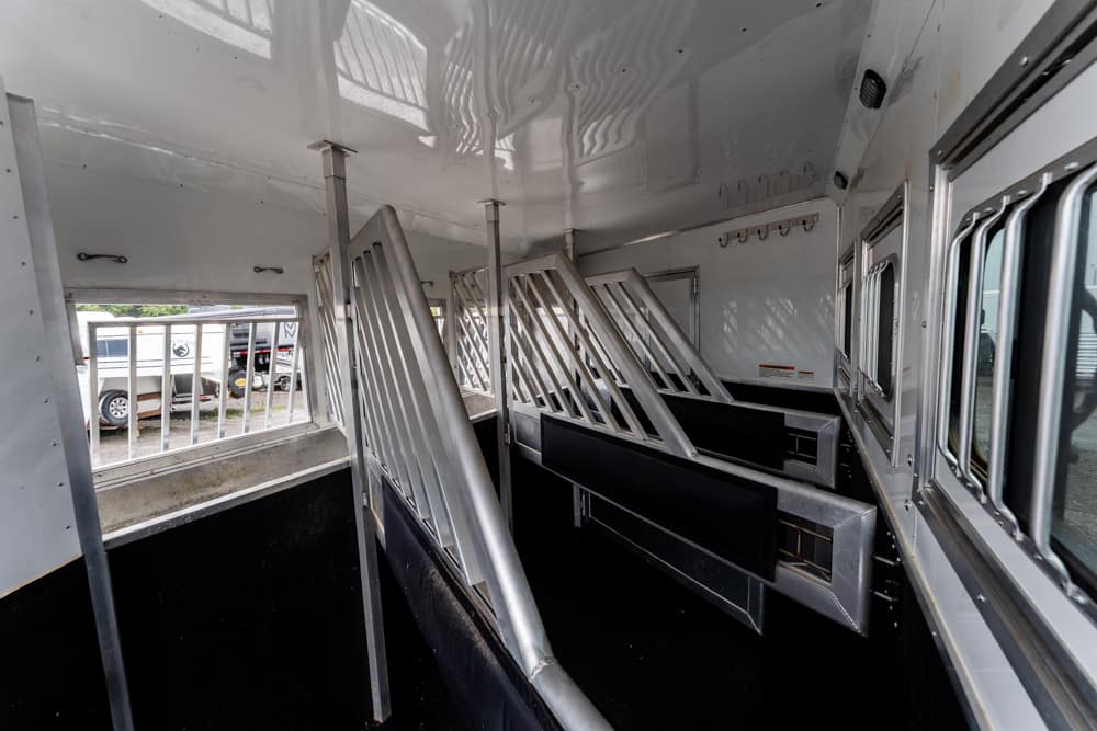 Interior of a white livestock trailer with metal stall dividers and black padded walls, windows along the side door area visible outside.