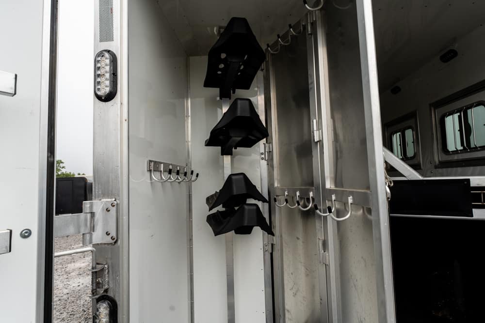Interior of a horse trailer with three black saddle racks mounted on a central rail and wall hooks nearby, outdoors visible through doorway.