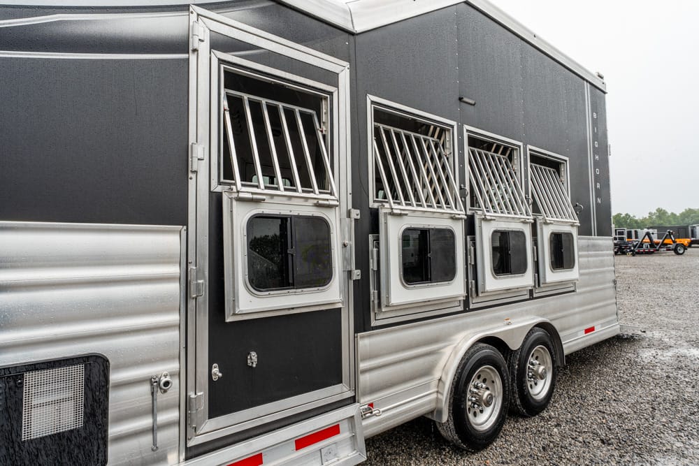 Gray horse trailer with ventilated stall windows and tandem wheels, parked on gravel surface in a lot.