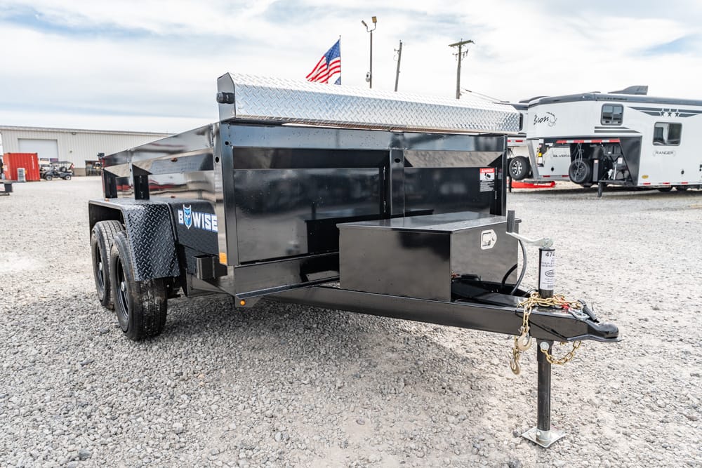 Black tandem-axle utility trailer with a diamond-plate toolbox on top, parked on a gravel lot, hitch and safety chains visible in front.
