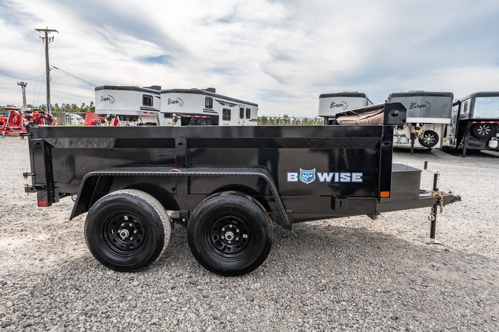Black dual-axle utility trailer with a Bowise logo parked on a gravel lot, surrounded by RVs and trucks in the background.