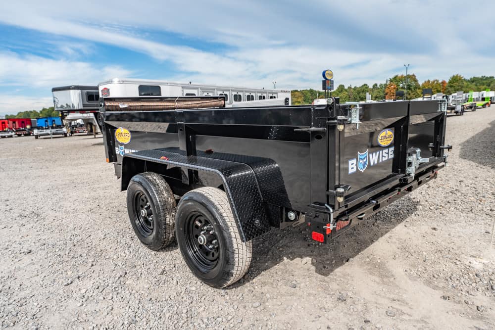Black dual-axle utility trailer with diamond-plate fenders in a gravel trailer yard, logo visible on the side.