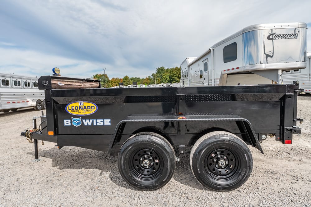 Black dual-axle dump trailer with Bowise and Leonard branding, parked on a gravel lot with RVs in the background; cargo bed sits level and shielded by a metal side rail.