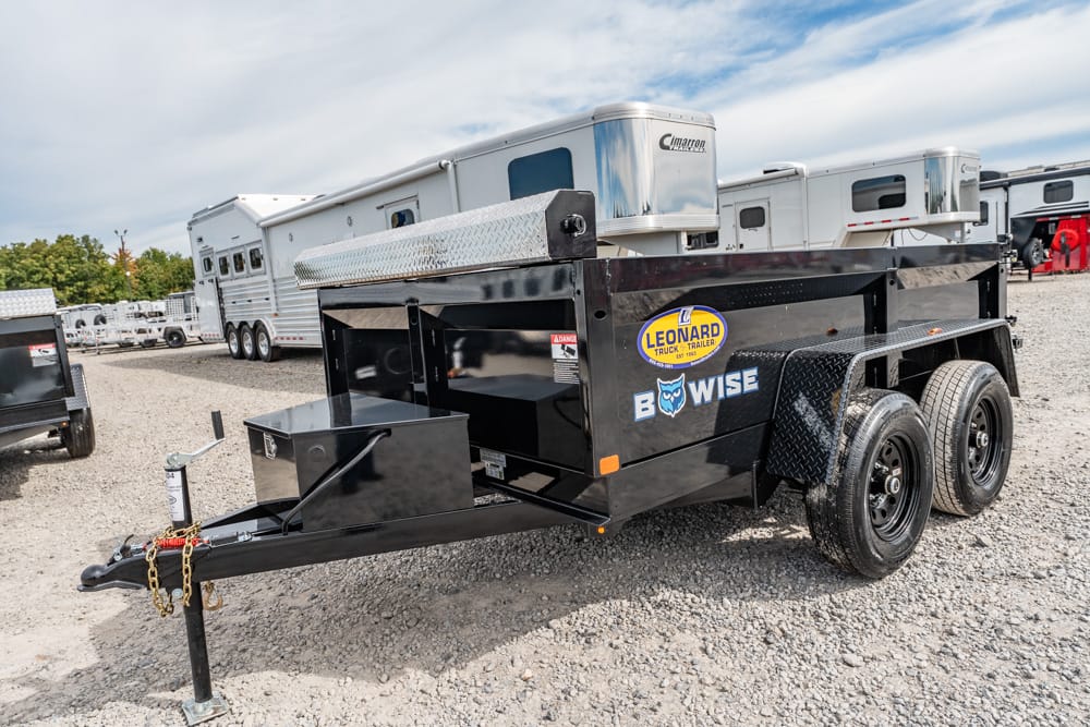 Black dual-axle BowWise utility trailer on a gravel lot with RVs in the background, featuring a diamond-plate toolbox at the front and a tow hitch with chains.