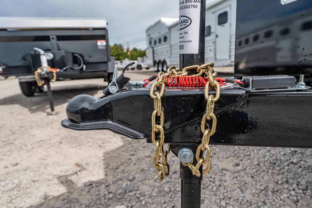 Close-up of a black trailer coupler with gold chains and a red safety coil securing it, in a gravel lot outside trailers.