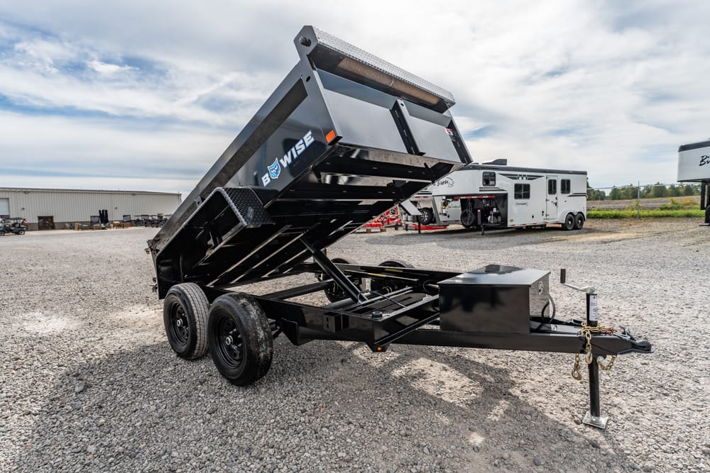 Black dual-axle dump trailer with its bed raised on a gravel lot, showing a hitch and storage box in the foreground.
