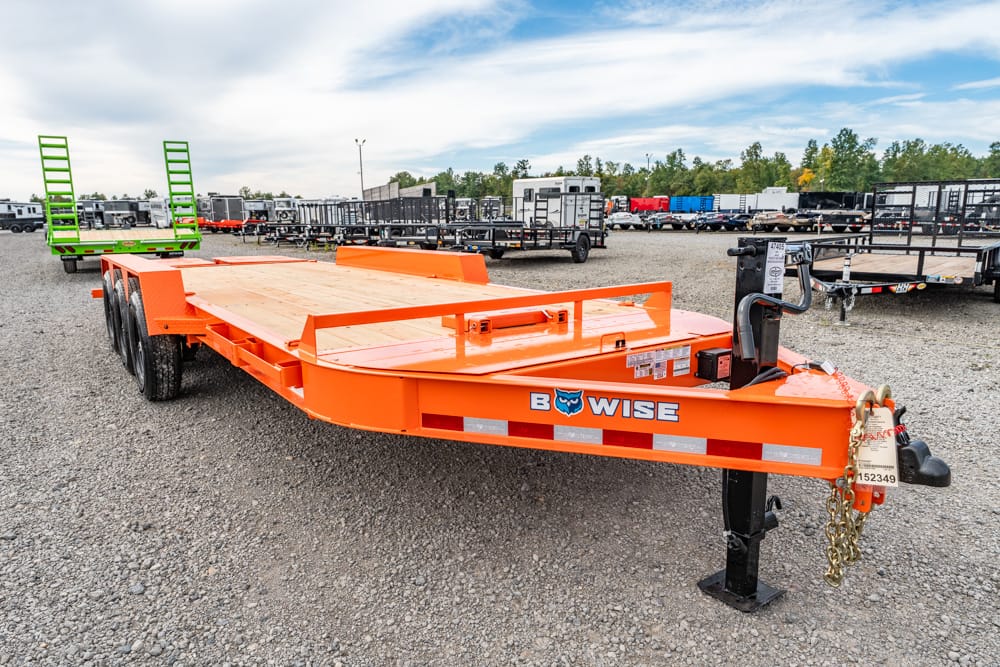 Orange B-WISE utility trailer with a flatbed and dual rear loading ramps in a gravel trailer yard.