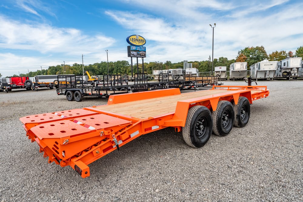 Bright orange multi-axle flatbed trailer with wooden deck in a gravel lot, ready for heavy hauling equipment or cars.