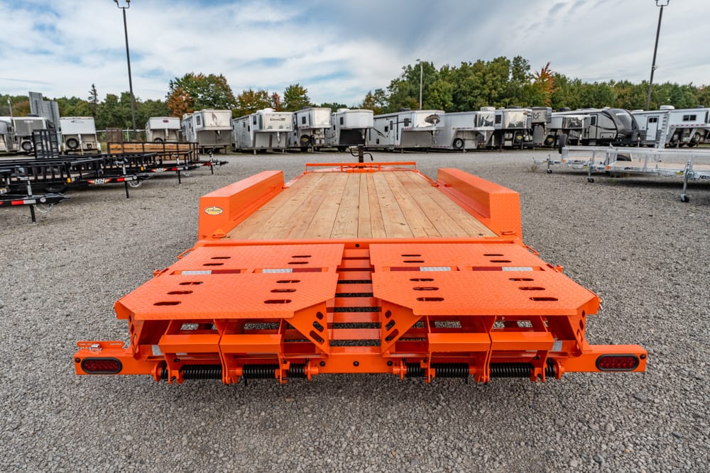 Orange heavy-duty flatbed trailer with rear ramps folded down in a gravel lot, surrounded by other trailers.