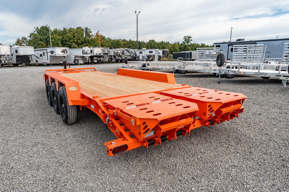 Orange multi-axle flatbed trailer with wooden deck, parked in a gravel trailer yard among other trailers, featuring ramp sections at the rear for loading