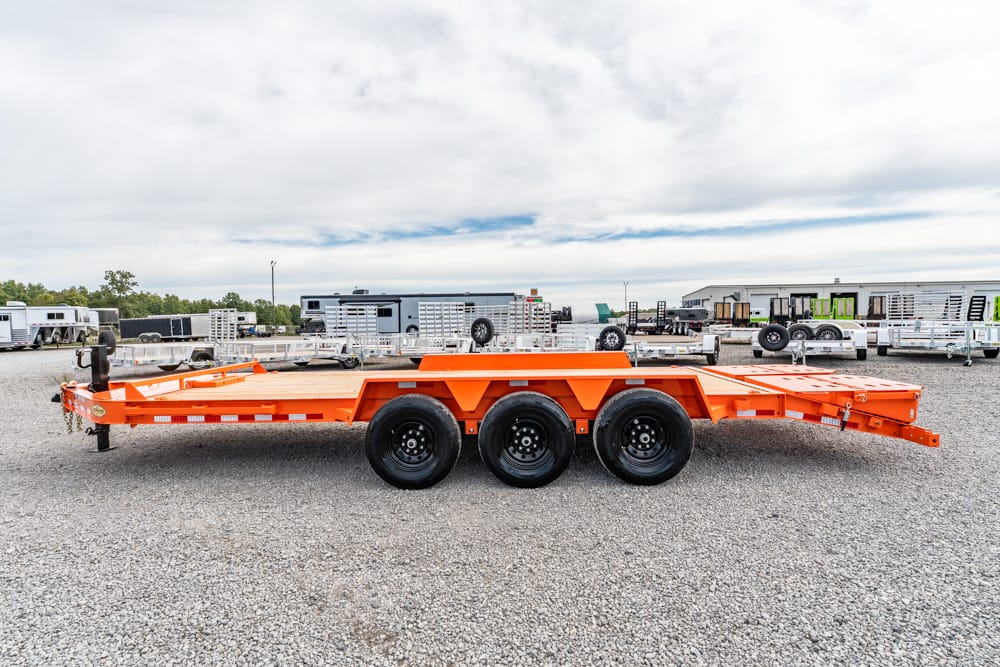 Orange heavy-duty trailer with three axles, parked on a gravel lot among other trailers and equipment, ready for hauling.