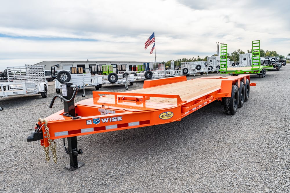 Bright orange heavy-duty trailer with a wooden deck sits in a gravel lot, other trailers visible in the background and an American flag overhead.
