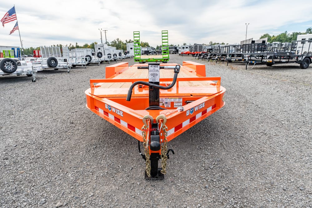 Orange utility trailer on a gravel lot, hitch with safety chains in the foreground, surrounded by rows of trailers and equipment with an American flag in the background