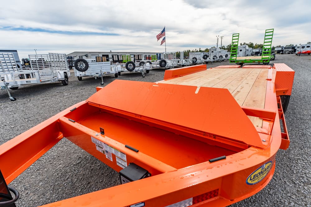 Orange utility trailer with a wooden deck and folding ramps in a dealership lot; American flag in the background.
