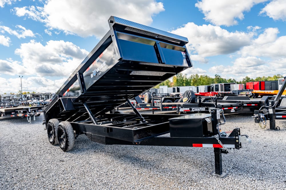 Black tilt-bed dump trailer raised on hydraulic supports in a gravel lot with other trailers nearby)