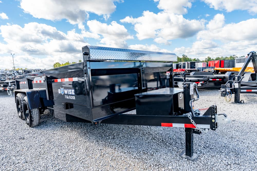 Black tandem-axle dump trailer on a gravel lot, with a diamond-plate toolbox at the front and safety chains attached.