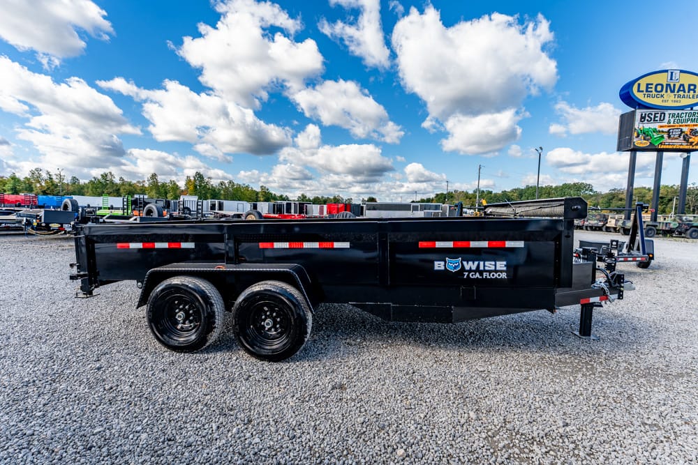 Black dual-axle dump trailer on a gravel lot, branded B-WISE 7 GA. floor with red reflective stripes. Vision includes clear blue sky and other trailers in the background.
