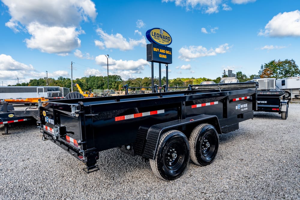 Black dual-axle dump trailer on a gravel lot, with other trailers and a Leonard sign visible in the background under a blue sky.