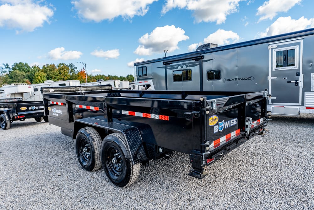 Black dual-axle dump trailer with reflective red/white tape on a gravel lot, BWISE branding, with a silver RV in the background.