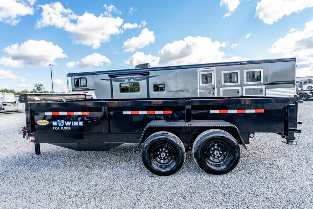 Side view of a black dual-axle dump trailer on a gravel lot, with a silver RV in the background, showing red-and-white reflective trim on its sides.