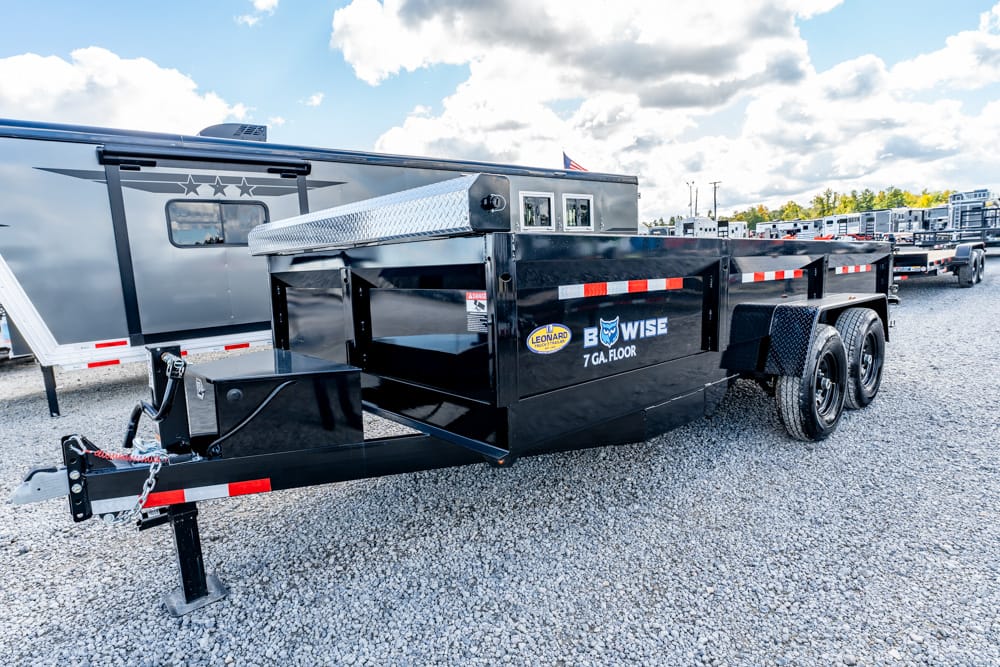 Black dual-axle utility trailer with a chrome diamond-plate toolbox at the front, parked on a gravel lot with other trailers nearby.