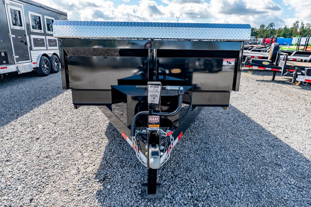 Front view of a black dump trailer on a gravel lot, with a diamond-plate toolbox and hitch area visible at the tongue.