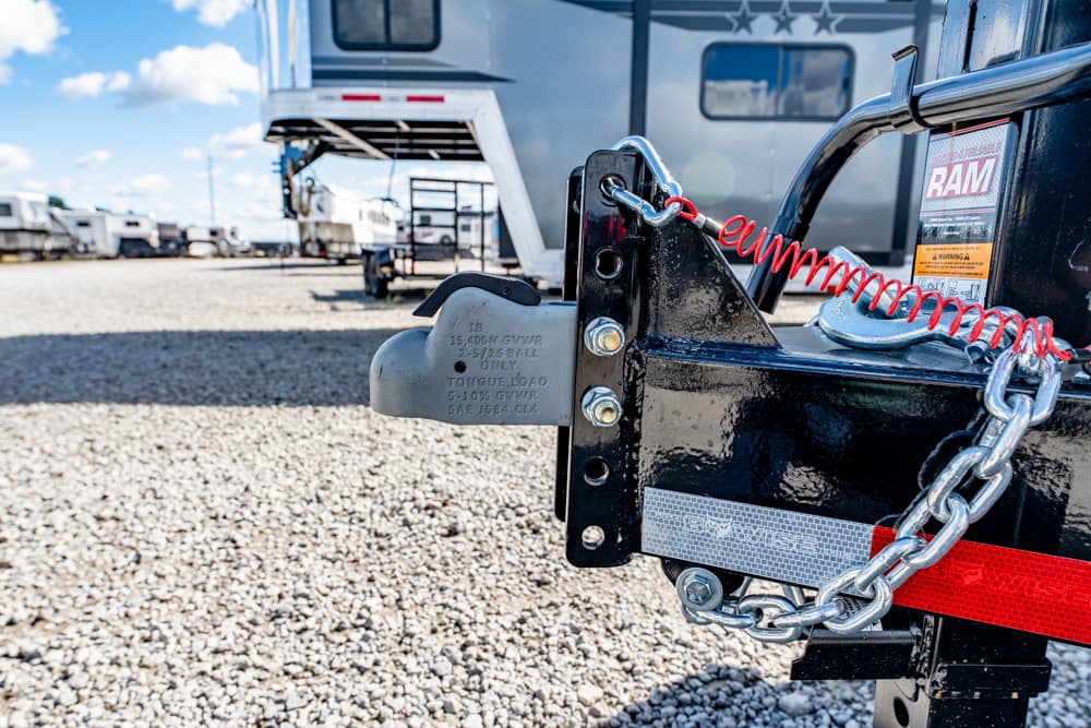 Close-up of a trailer hitch lock and metal chain on a black trailer frame, with a red reflective strip and several RVs in the gravel lot.