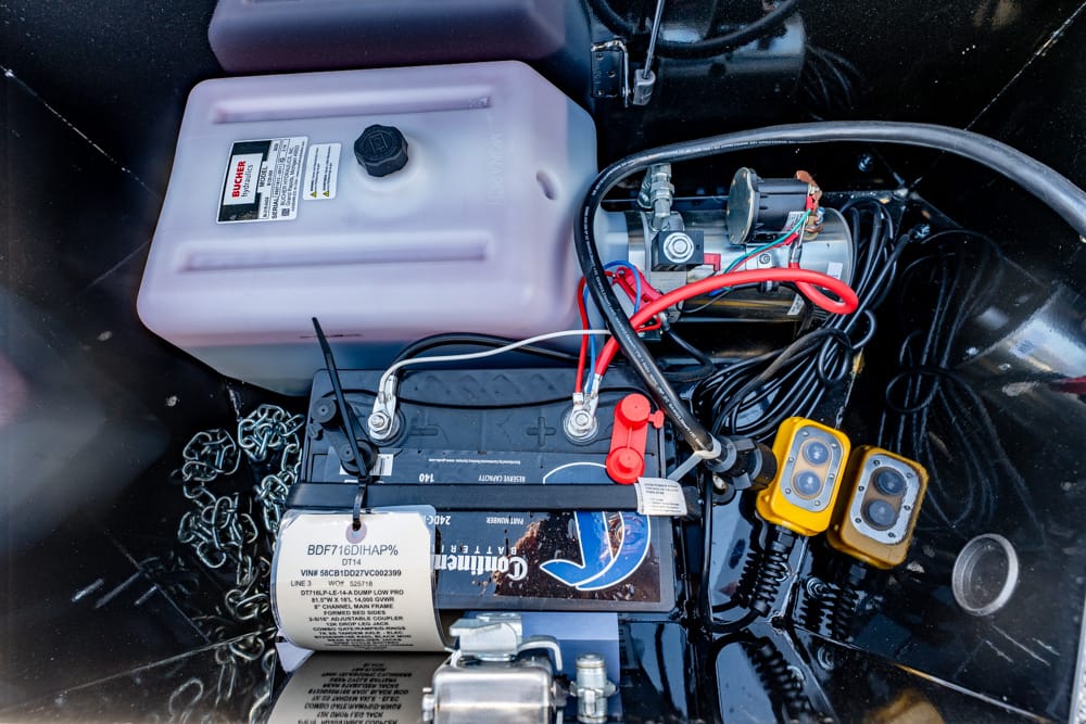 Boat battery bay with a large black battery, red/black cables, a pink translucent reservoir, and two yellow handheld meters nearby.