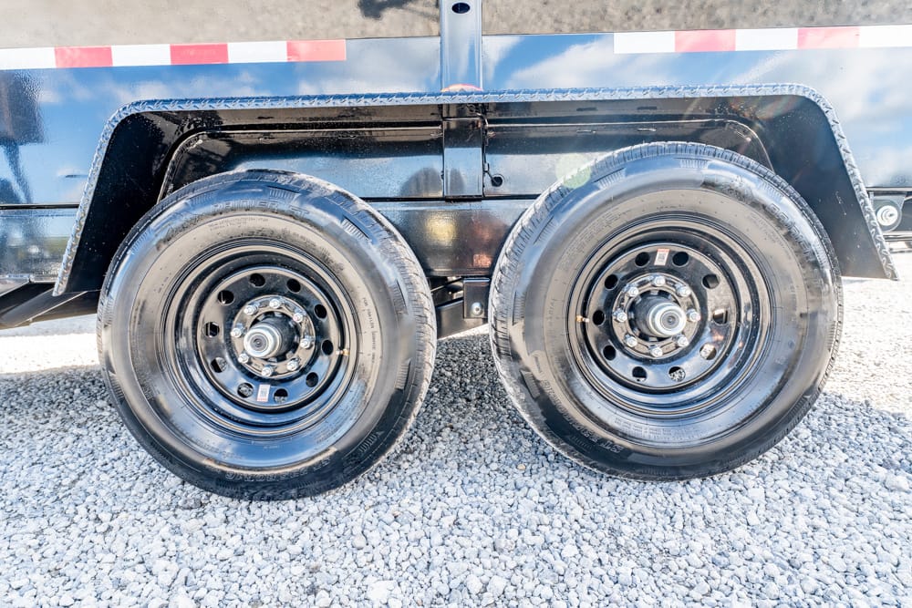 Dual black trailer wheels with hubs on a gravel surface under a fender and reflective tape.