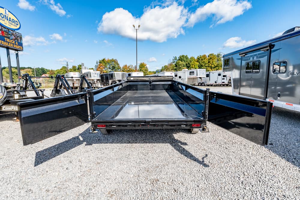 Rear view of a black utility trailer with its side ramps extended, resting on a gravel lot with other trailers and RVs nearby.