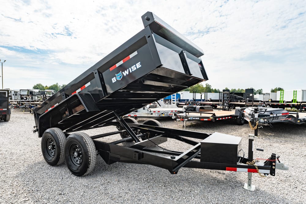 Black dual-axle hydraulic dump trailer raised for dumping in a gravel lot with other trailers in the background, Wise logo visible on the bed