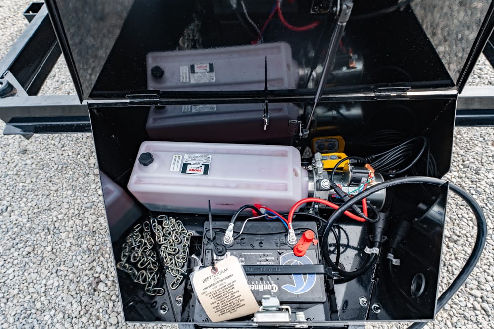 Open storage compartment in a trailer showing a car battery, red/black cables, a white plastic reservoir, and chains inside.