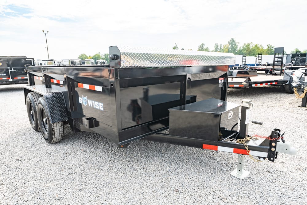 Black tandem-axle utility/dump trailer with diamond-plate toolbox, parked on gravel; hitch and safety chains in view.