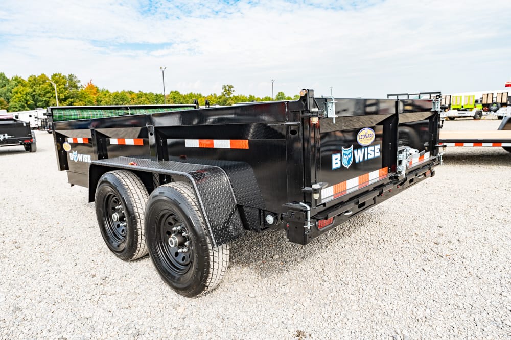 Black two-axle dump trailer with orange reflective stripes parked on a gravel lot.