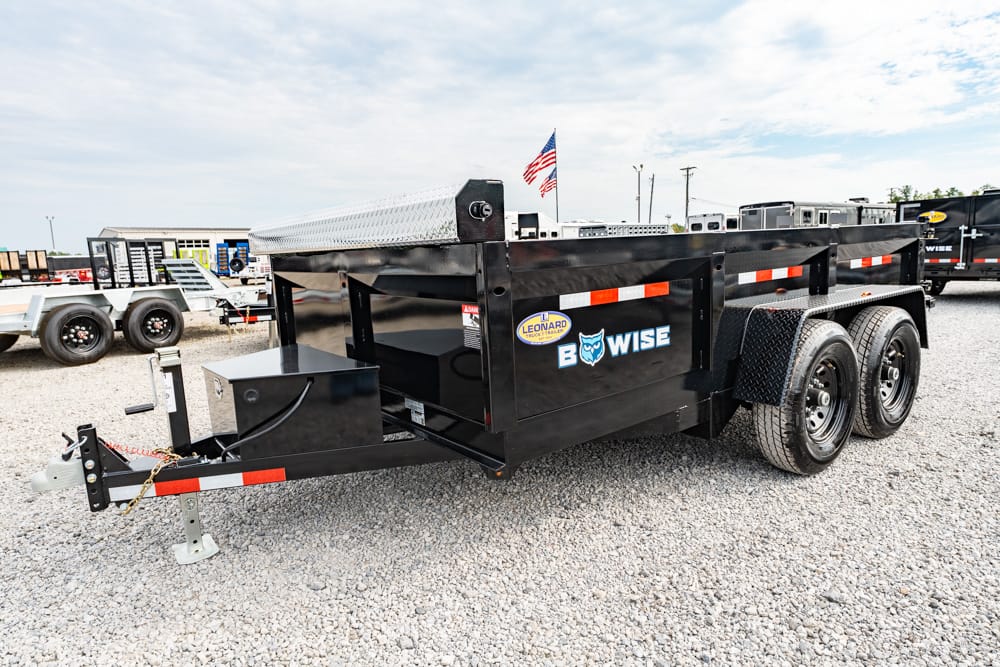 Black tandem-axle dump trailer with reflective orange/white stripes, parked on a gravel lot, showing the B-WISE logo and an American flag on top.