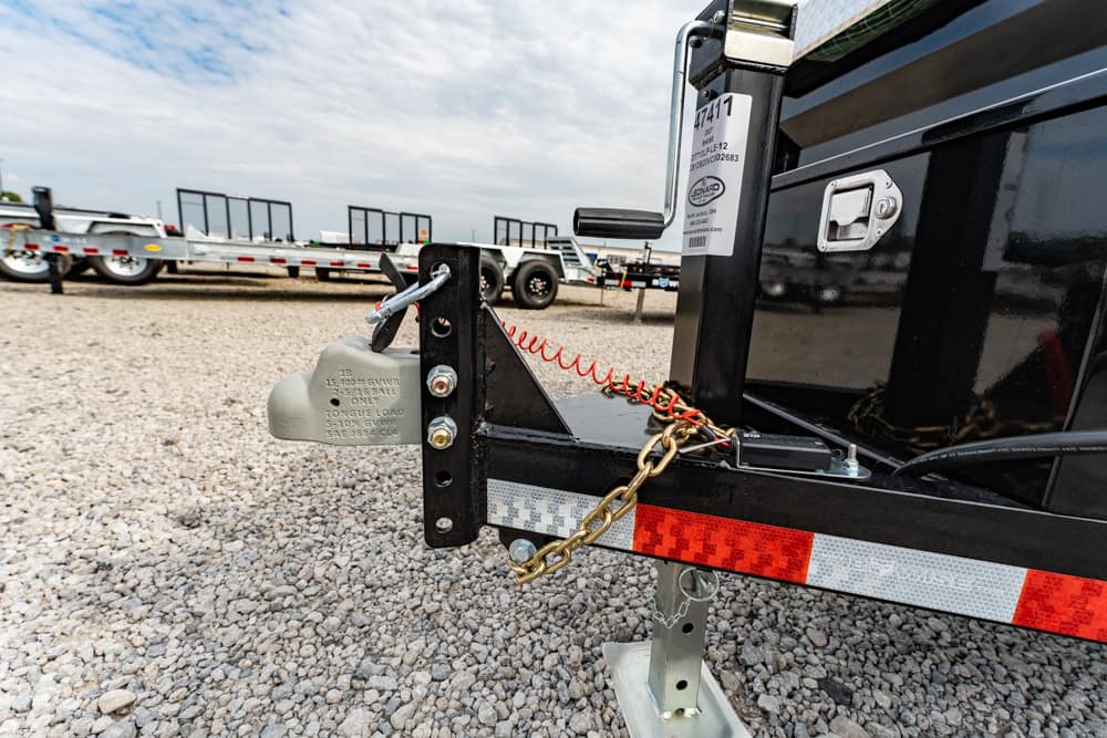 Close-up of a trailer hitch with safety chains attached to a black trailer on a gravel lot, with other trailers in the background.