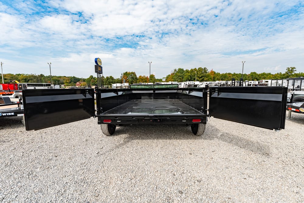 Black flatbed utility trailer with side gates swung open, parked on a gravel lot with other trailers and trucks in the background.