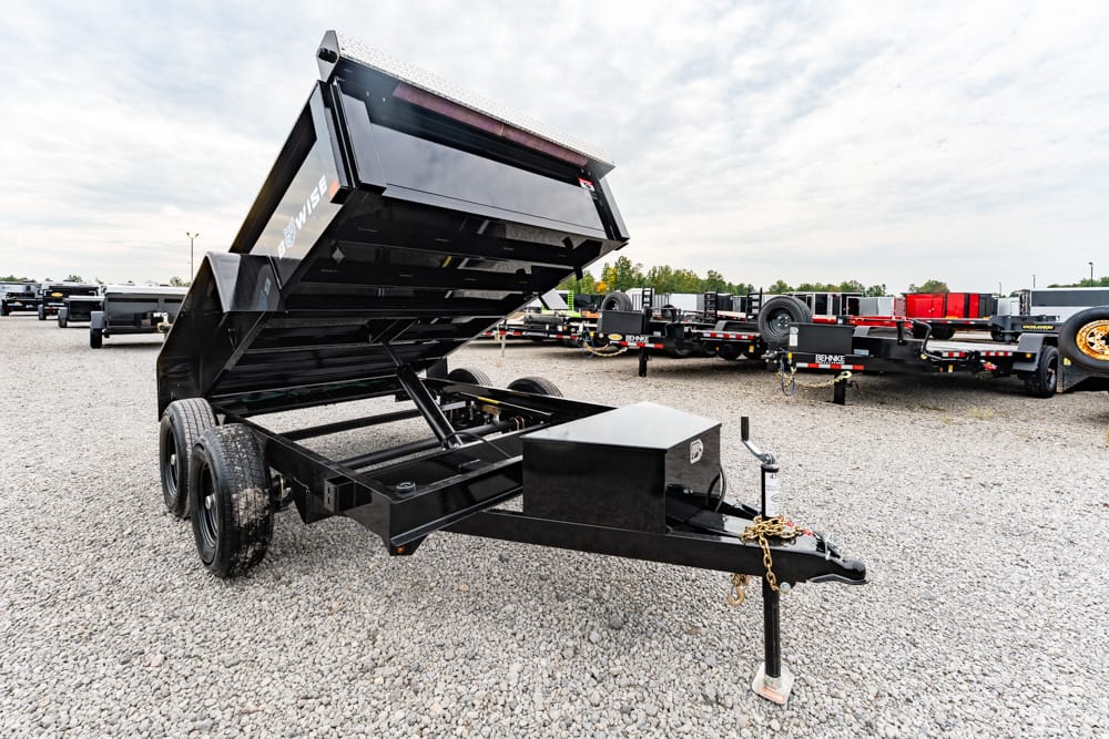 Black hydraulic-dump trailer with bed raised, in a gravel lot with other trailers in the background.