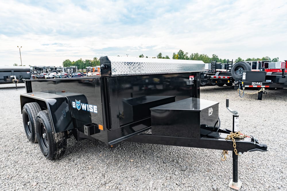 Black utility trailer with dual axles, a diamond‑plate toolbox, and a front hitch on a gravel lot, labeled B‑WISE.