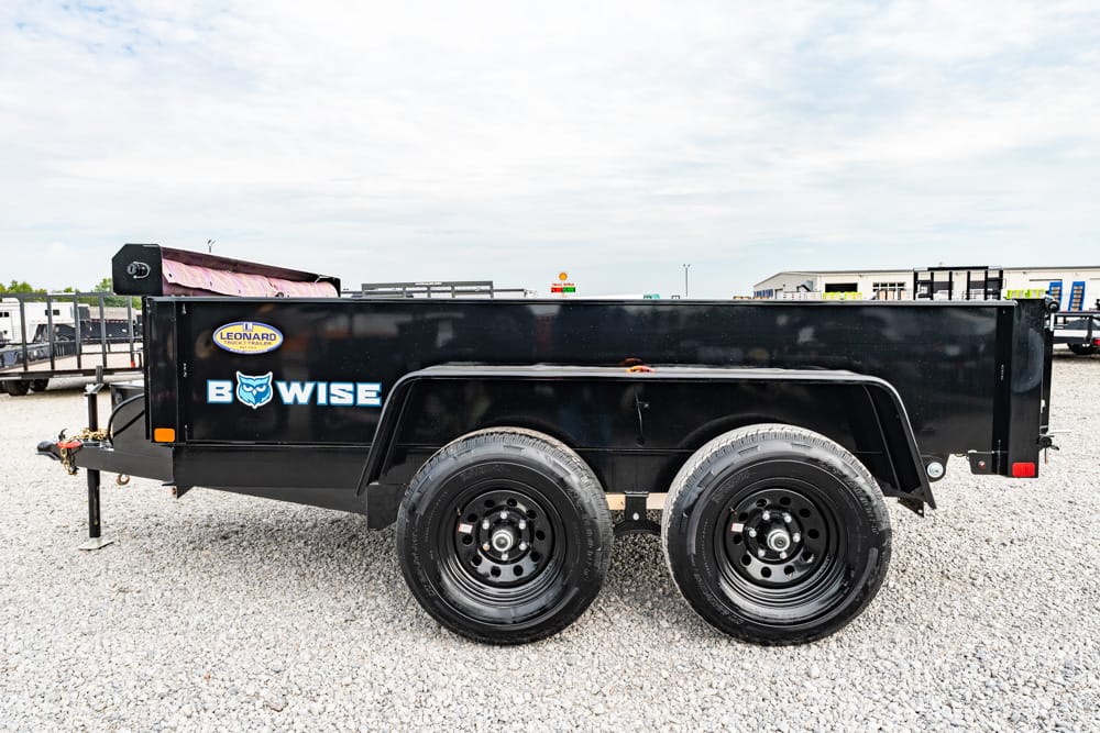 Black dual-axle utility trailer with BowWise logo and owl icon, parked on a gravel lot with industrial buildings in the background.
