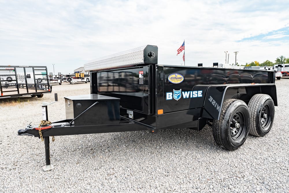 Black dual-axle BowWise utility dump trailer with a diamond-plate toolbox at the front, parked on a gravel lot with an American flag in the background.